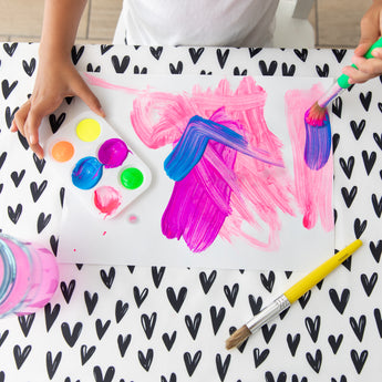 A child paints with brushes on paper at a table covered with a Bumkins Splat Mat: Hearts, with a paint palette nearby.