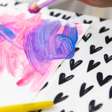 Close-up of a child painting with pink and purple on white paper over a Bumkins Splat Mat: Hearts.