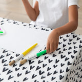 A child in a white shirt sits at a table covered with a Bumkins Splat Mat: Hearts, holding green and yellow brushes next to paper.