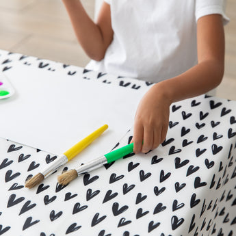 A child in a white shirt sits at a table covered with a Bumkins Splat Mat: Hearts, holding green and yellow brushes next to paper.