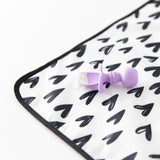 Close-up of a purple baby spoon with food sitting on a Bumkins Splat Mat: Hearts, made from easy-wipe fabric, on a white background.