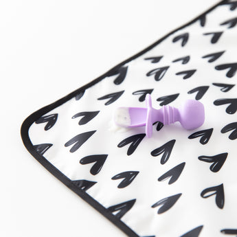 Close-up of a purple baby spoon with food sitting on a Bumkins Splat Mat: Hearts, made from easy-wipe fabric, on a white background.