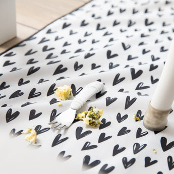 Close-up of a white baby fork with food bits on an easy-clean Bumkins Splat Mat: Hearts.