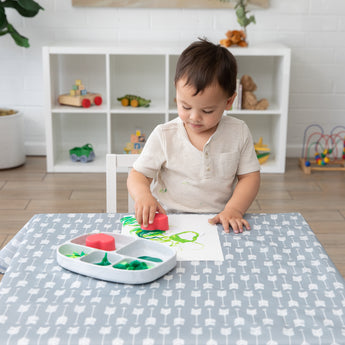 A boy uses Bumkins Silicone Grip Dish with Lid (5 Section): Marble to paint at a cloth-covered table in a bright playroom.
