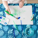 A child paints with green, blue, brushes, and a water cup on white paper using Bumkins Splat Mat: Blue Tropic for easy cleanup.