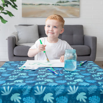 Young child painting on a Bumkins Splat Mat: Blue Tropic, smiling and looking up—easy cleanup made simple.