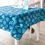 On a white table, a person enjoys a blue drink with Bumkins Splat Mat: Blue Tropic, appreciating its easy-cleaning, waterproof fabric.