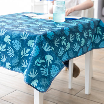 On a white table, a person enjoys a blue drink with Bumkins Splat Mat: Blue Tropic, appreciating its easy-cleaning, waterproof fabric.