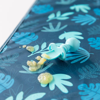 Baby food spills from a blue spoon on a Bumkins Splat Mat: Blue Tropic with easy-clean leaf designs.
