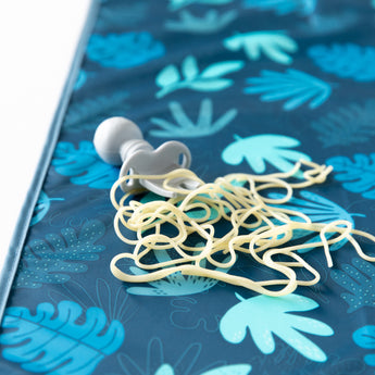 A tangled pacifier clip with rubber bands rests on the Bumkins Splat Mat: Blue Tropic, a waterproof fabric thats easy to clean.
