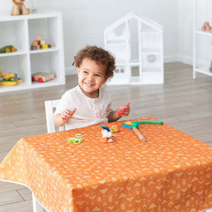 A child smiling at table with craft supplies on a table covered in a Bumkins Splat Mat: Grounded, in a bright room with toys on shelves.