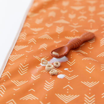 Close-up of small cereal pieces and spilled milk next to a brown spoon on Bumkins Splat Mat: Grounded on a white background.
