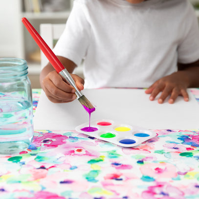 A child in a white shirt paints with a purple brush on a blank sheet. Nearby are water jars and a palette with red, purple, blue, yellow, and green paint. The table is covered with a colorful, abstract floral design.
