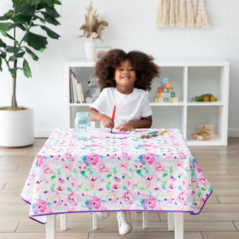 A child smiles while painting on a table covered with a Bumkins Splat Mat: Watercolor. A bookshelf and plant add charm to the background.