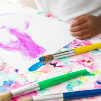 A child paints with bright colors, surrounded by brushes, on a stain-resistant Bumkins Splat Mat: Watercolor.