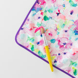 Close-up of Bumkins Splat Mat: Watercolor, colorful and stain-resistant, on a white background with a paint brush and paint smear on top.