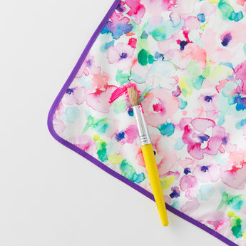 Close-up of Bumkins Splat Mat: Watercolor, colorful and stain-resistant, on a white background with a paint brush and paint smear on top.