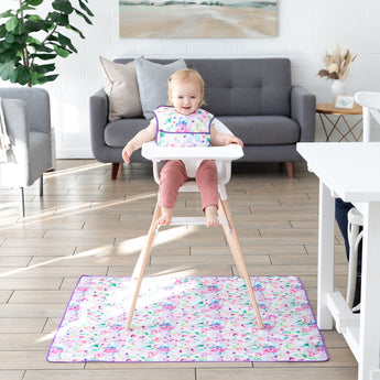A smiling toddler in a high chair wearing a floral bib sits on a Bumkins Splat Mat: Watercolor, featuring stain-resistant fabric.