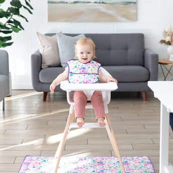 A smiling baby sits in a high chair with a colorful bib on a Bumkins Splat Mat: Watercolor, near a gray sofa and green plant.