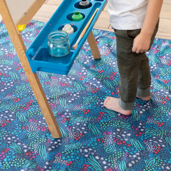 A child stands barefoot on a Bumkins Splat Mat: Animal Prints next to a painting easel with jars of paint.