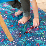 Close-up of a child reaching for a paintbrush on a Bumkins Splat Mat: Animal Prints, made of waterproof fabric for easy cleaning.