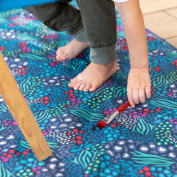 Close-up of a child reaching for a paintbrush on a Bumkins Splat Mat: Animal Prints, made of waterproof fabric for easy cleaning.