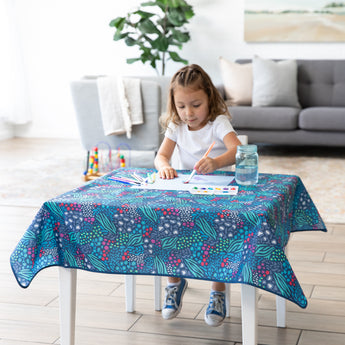 A child paints at a table covered with a Bumkins Splat Mat: Animal Prints; a plant and couch line the background of the bright room.