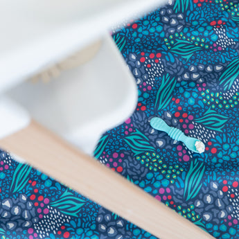 A silicone spoon with food rests on a Bumkins Splat Mat: Animal Prints underneath a high chair.