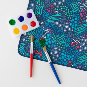 Two paintbrushes with red and blue handles sit on a Bumkins Splat Mat: Animal Prints near a palette of paints on a white background.