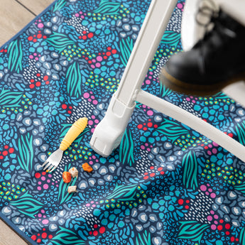 Bumkins Splat Mat: Animal Prints lays under a high chair, with a yellow fork and pieces of food on top.