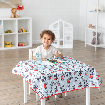 Child painting on a Bumkins Splat Mat: Mickey Mouse, at a table in a bright room with toys on shelves.