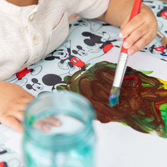 A child paints brown and green on paper, protected by a Bumkins Splat Mat: Mickey Mouse-themed tablecloth underneath.