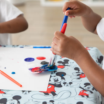 Child painting on a Mickey Mouse Splat Mat by Bumkins, crafted from waterproof and machine washable fabric.