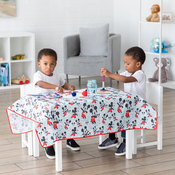 Two kids joyfully paint at a small table covered with the machine-washable Bumkins Splat Mat: Mickey Mouse in their cozy room.