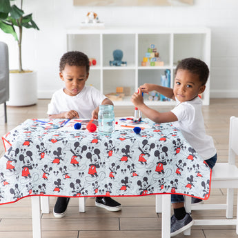 Two young children enjoy arts and crafts on a Bumkins Splat Mat: Mickey Mouse design, machine washable, in a bright room.