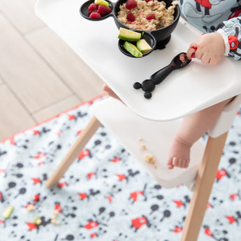 Child in high chair eating oatmeal, raspberries, cucumber with a Mickey Mouse spoon on Bumkins machine-washable Splat Mat: Mickey Mouse.