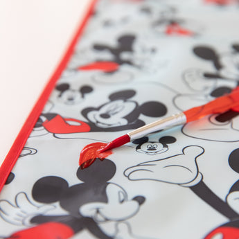 Machine washable waterproof splat mat by Bumkins featuring a red paintbrush design and Mickey Mouse cartoon faces.