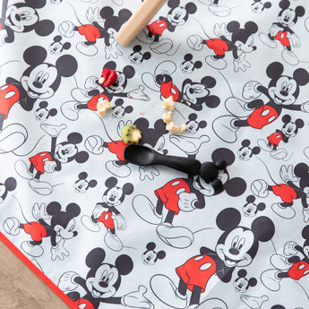 Bumkins Mickey Mouse Splat Mat features scattered food, a black spoon, and rolling pin on waterproof fabric. Machine washable for easy cleanup.