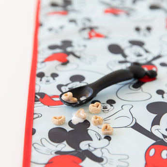 A black spoon with cereal and milk rests on the waterproof, machine washable Bumkins Splat Mat featuring Mickey Mouse.