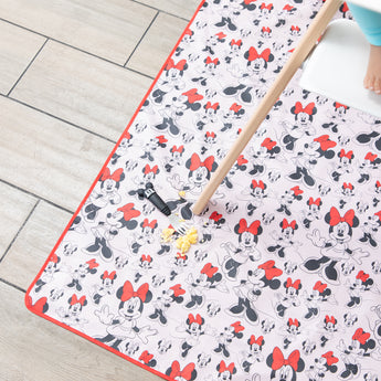 Scrambled eggs and a Mickey Mouse fork sit on a Bumkins Splat Mat: Minnie Mouse, under a wooden highchair leg on the floor. 