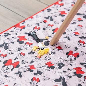 Scrambled eggs and a Mickey Mouse fork sit on a Bumkins Splat Mat: Minnie Mouse, under a wooden table leg on the floor. 