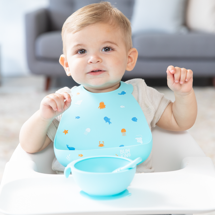A child wearing a blue silicone bib sits in a high chair with a blue bowl on the tray with a gray couch in the background.
