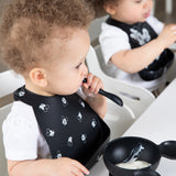 Two toddlers in Bumkins Silicone Bib 2 Pack: Disney Mickey and Minnie Mouse eat yogurt with black spoons and bowls at a white table.