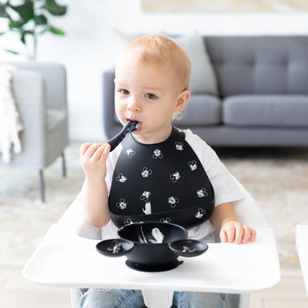 A toddler in a high chair enjoys baby-led weaning with Bumkins Silicone Feeding 3-Piece Set: Mickey Mouse Faces B+W in a bright living room.