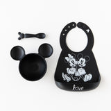 Bumkins Silicone Bib: Mickey + Minnie Mouse lays next to a black Mickey Mouse-shaped bowl & spoon set on a white background.