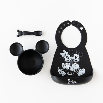 Bumkins Silicone Bib: Mickey + Minnie Mouse lays next to a black Mickey Mouse-shaped bowl & spoon set on a white background.