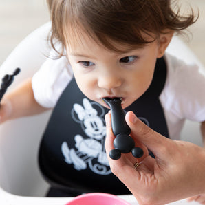 Close-up of a hand feeding a child wearing a black bib in a high chair with a Bumkins Silicone Dipping Spoons: Minnie Mouse (Black and Pink).