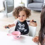 A curly haired child holds Bumkins Silicone Dipping Spoons: Minnie Mouse (Black and Pink) over a pink Minnie Mouse-shaped bowl of food.
