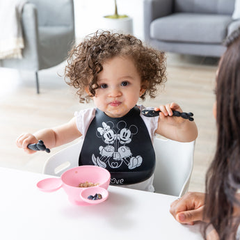 A curly haired child holds Bumkins Silicone Dipping Spoons: Minnie Mouse (Black and Pink) over a pink Minnie Mouse-shaped bowl of food.