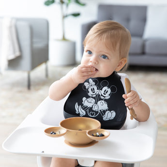 A baby in a high chair wears a Bumkins Silicone Bib 2 Pack: Disney Mickey and Minnie Mouse, eating from a gold bowl and holding a spoon.